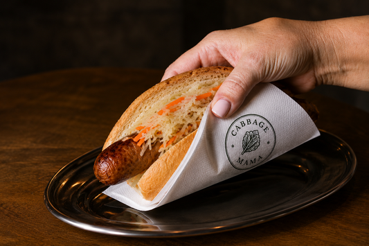 Grilled sausage sandwich topped with Cabbage Mama sauerkraut and carrot, wrapped in a Cabbage Mama napkin.